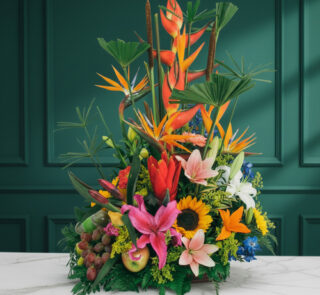 Arreglo floral tropical con heliconias, aves del paraíso, lirios, girasoles y frutas variadas sobre una mesa de mármol blanca frente a pared verde oscura
