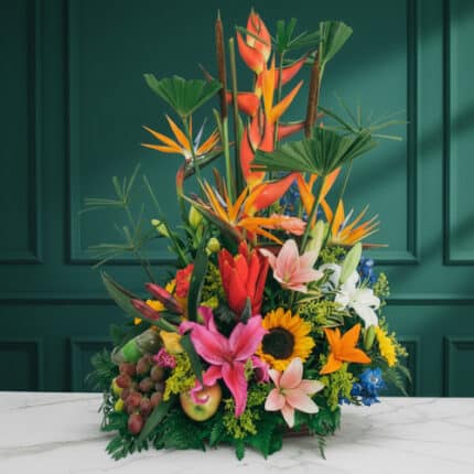 Arreglo floral tropical con heliconias, aves del paraíso, lirios, girasoles y frutas variadas sobre una mesa de mármol blanca frente a pared verde oscura