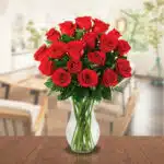 Arreglo de rosas rojas frescas en un jarrón de cristal transparente colocado sobre una mesa de madera, con fondo de sala desenfocado