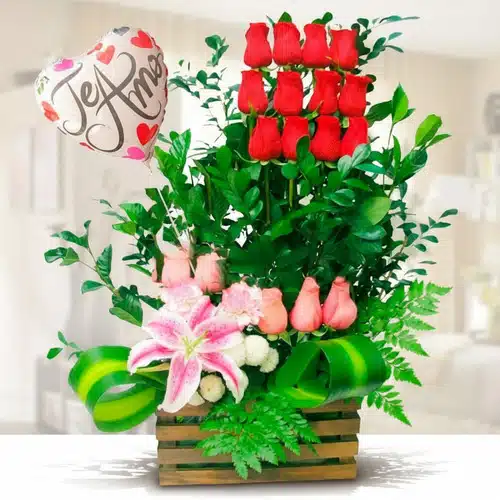 Arreglo floral en base de madera con rosas rojas verticales, rosas rosadas, lirio rosa, follaje verde y globo metálico en forma de corazón con la frase Te amo