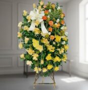 corona funeraria ovalada de rosas amarillas y naranjas con lazo blanco sobre caballete de madera en interior