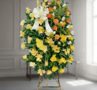 corona funeraria ovalada de rosas amarillas y naranjas con lazo blanco sobre caballete de madera en interior