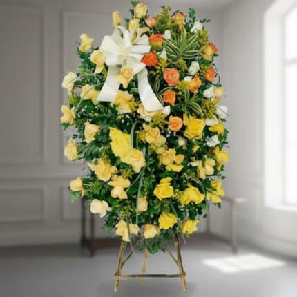 corona funeraria ovalada de rosas amarillas y naranjas con lazo blanco sobre caballete de madera en interior