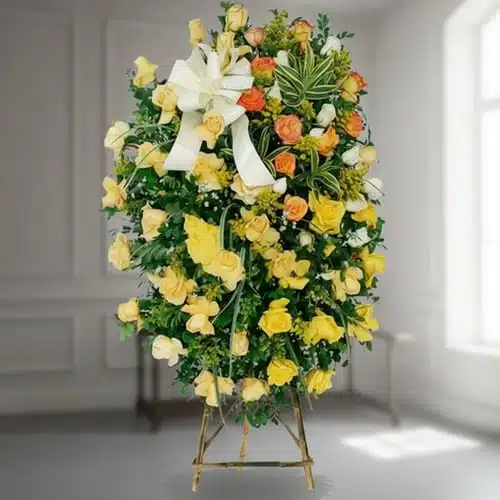 corona funeraria ovalada de rosas amarillas y naranjas con lazo blanco sobre caballete de madera en interior