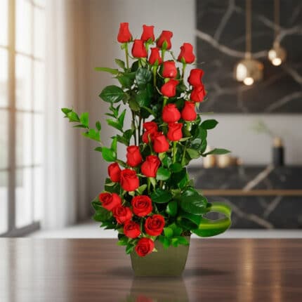 Arreglo floral alto de rosas rojas con follaje verde en florero cuadrado verde sobre mesa de madera en sala moderna desenfocada