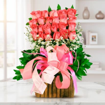 arreglo de rosas rosadas en dos niveles con follaje verde, flores blancas pequeñas y lazos rosados sobre base de madera, colocado sobre mesa blanca en interior