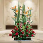 Arreglo floral grande con rosas rojas, aves del paraíso naranjas y lirios rosados sobre mesa de mármol en interior elegante