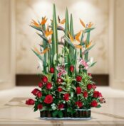 Arreglo floral grande con rosas rojas, aves del paraíso naranjas y lirios rosados sobre mesa de mármol en interior elegante