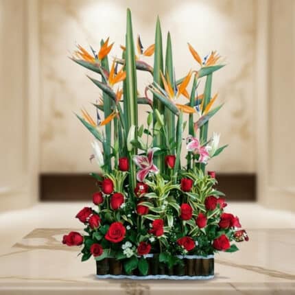 Arreglo floral grande con rosas rojas, aves del paraíso naranjas y lirios rosados sobre mesa de mármol en interior elegante