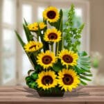 Arreglo de girasoles amarillos con centro marrón y follaje verde en maceta rectangular sobre mesa de madera