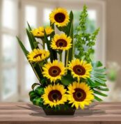 Arreglo de girasoles amarillos con centro marrón y follaje verde en maceta rectangular sobre mesa de madera