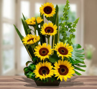 Arreglo de girasoles amarillos con centro marrón y follaje verde en maceta rectangular sobre mesa de madera