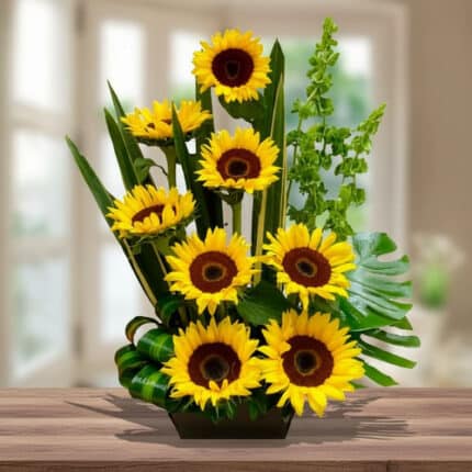 Arreglo de girasoles amarillos con centro marrón y follaje verde en maceta rectangular sobre mesa de madera