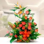 arreglo floral con rosas naranjas, flores ave del paraíso y follaje verde sobre base de madera