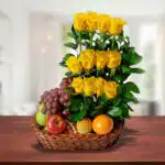 Canasta de mimbre sobre mesa de madera con uvas, manzanas, naranja y un arreglo vertical de rosas amarillas