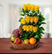 Canasta de mimbre sobre mesa de madera con uvas, manzanas, naranja y un arreglo vertical de rosas amarillas