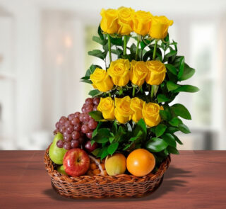 Canasta de mimbre sobre mesa de madera con uvas, manzanas, naranja y un arreglo vertical de rosas amarillas