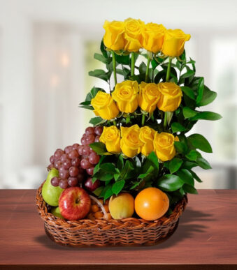 Canasta de mimbre sobre mesa de madera con uvas, manzanas, naranja y un arreglo vertical de rosas amarillas