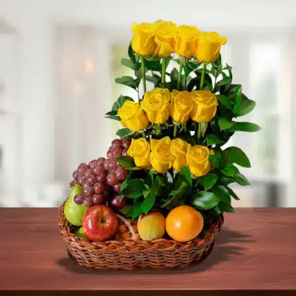 Canasta de mimbre sobre mesa de madera con uvas, manzanas, naranja y un arreglo vertical de rosas amarillas