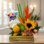 Arreglo de frutas variadas con girasol, heliconias y globo metálico con texto Feliz Día sobre base de bambú en interior iluminado