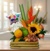 Arreglo de frutas variadas con girasol, heliconias y globo metálico con texto Feliz Día sobre base de bambú en interior iluminado