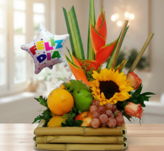 Arreglo de frutas variadas con girasol, heliconias y globo metálico con texto Feliz Día sobre base de bambú en interior iluminado
