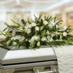 Arreglo fúnebre de flores blancas y follaje verde colocado sobre un ataúd metálico en una sala iluminada