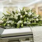 Arreglo fúnebre de flores blancas y follaje verde colocado sobre un ataúd metálico en una sala iluminada