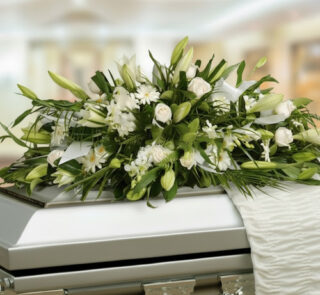 Arreglo fúnebre de flores blancas y follaje verde colocado sobre un ataúd metálico en una sala iluminada