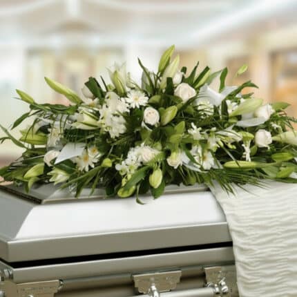 Arreglo fúnebre de flores blancas y follaje verde colocado sobre un ataúd metálico en una sala iluminada