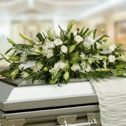 Arreglo fúnebre de flores blancas y follaje verde colocado sobre un ataúd metálico en una sala iluminada