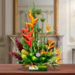 Arreglo floral tropical con heliconias naranjas, follaje verde y una botella de cerveza en el centro, sobre mesa de madera en interior elegante