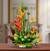 Arreglo floral tropical con heliconias naranjas, follaje verde y una botella de cerveza en el centro, sobre mesa de madera en interior elegante