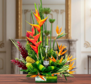 Arreglo floral tropical con heliconias naranjas, follaje verde y una botella de cerveza en el centro, sobre mesa de madera en interior elegante