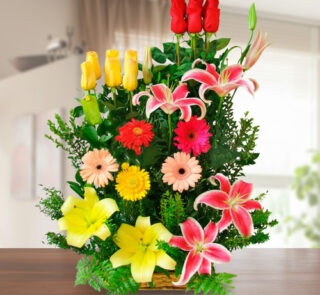 Arreglo floral vertical sobre mesa de madera con rosas rojas y amarillas, lirios rosados y amarillos y gerberas de varios colores sobre fondo de sala desenfocada