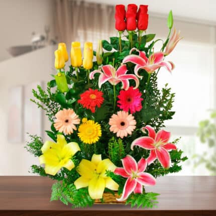 Arreglo floral vertical sobre mesa de madera con rosas rojas y amarillas, lirios rosados y amarillos y gerberas de varios colores sobre fondo de sala desenfocada