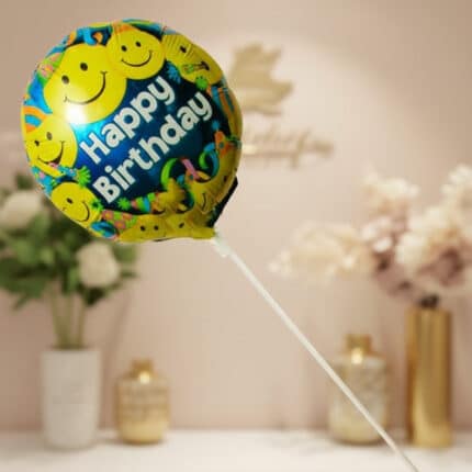 Globo redondo de cumpleaños con caritas felices amarillas y texto Happy Birthday sobre fondo colorido, sostenido por una varilla blanca en una sala decorada con flores y jarrones desenfocados