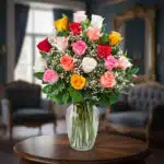 Arreglo floral de rosas rojas, rosas, blancas, amarillas y naranjas con paniculata en un jarrón de cristal sobre una mesa de madera en una sala elegante