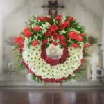 Corona fúnebre redonda con rosas rojas y flores blancas sobre caballete de madera en interior de iglesia desenfocada