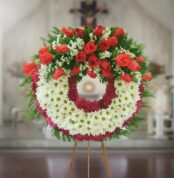 Corona fúnebre redonda con rosas rojas y flores blancas sobre caballete de madera en interior de iglesia desenfocada