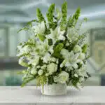 Arreglo de flores blancas con lirios, rosas y follaje verde en una canasta blanca sobre mesa de mármol