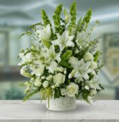 Arreglo de flores blancas con lirios, rosas y follaje verde en una canasta blanca sobre mesa de mármol