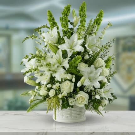 Arreglo de flores blancas con lirios, rosas y follaje verde en una canasta blanca sobre mesa de mármol