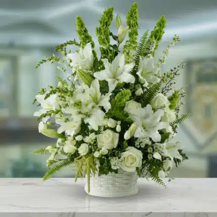Arreglo de flores blancas con lirios, rosas y follaje verde en una canasta blanca sobre mesa de mármol