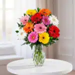 Ramo de gerberas rojas, rosas, naranjas, amarillas y blancas en un jarrón de cristal transparente sobre una mesa redonda blanca
