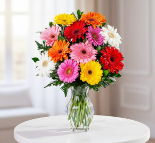 Ramo de gerberas rojas, rosas, naranjas, amarillas y blancas en un jarrón de cristal transparente sobre una mesa redonda blanca
