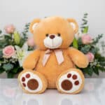 Oso de peluche color beige con lazo rosa sentado sobre superficie blanca, con flores rosadas y blancas de fondo