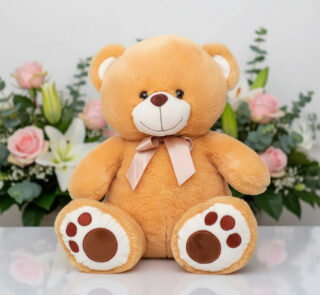 Oso de peluche color beige con lazo rosa sentado sobre superficie blanca, con flores rosadas y blancas de fondo