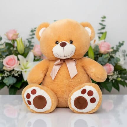 Oso de peluche color beige con lazo rosa sentado sobre superficie blanca, con flores rosadas y blancas de fondo