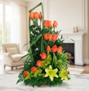 Arreglo floral vertical con rosas naranjas, lirios amarillos y follaje verde colocado sobre una mesa de mármol en una sala luminosa